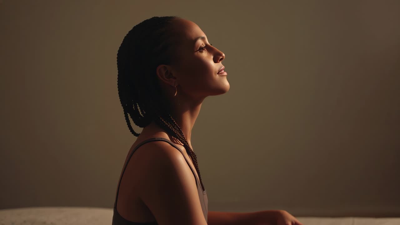 Inhaling woman lifting chin and smiling, reflecting in studio, braided hair, warm light, dark tank