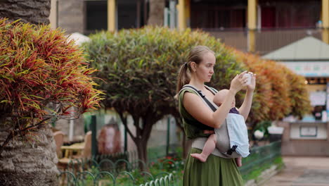 A-young-woman-travels-with-her-baby-in-a-kangaroo-backpack,-capturing-moments-on-her-mobile-phone.-As-she-strolls,-she-gazes-at-the-phone-screen