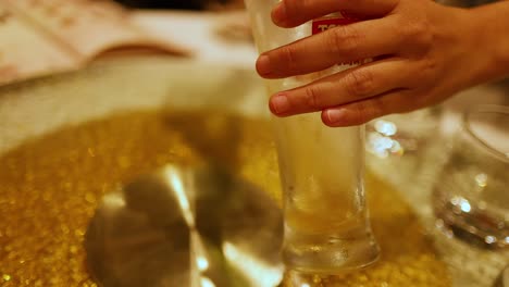 a hand pours beer into a glass on a glittering table in a lively bar setting, capturing a casual, social atmosphere