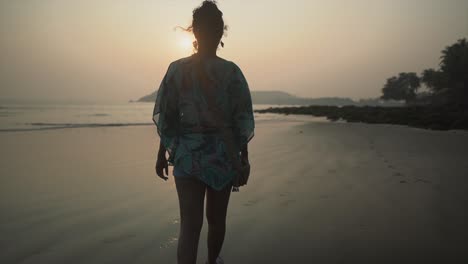 landscape view of a young woman walking on a sandy tropical beach by the ocean, towards the sunset