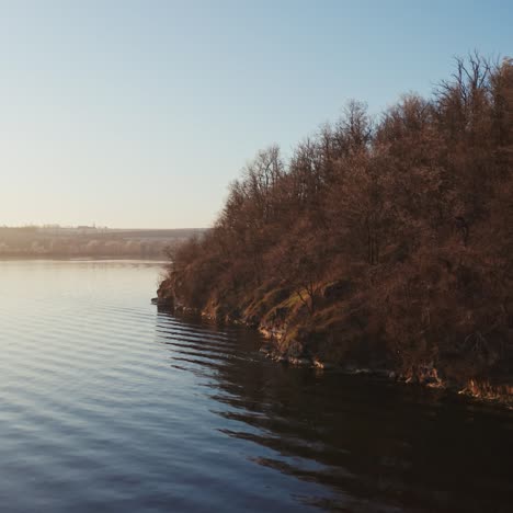 Flying over the water. Trees on the hill near the water in winter season. River in the evening. Drone view.