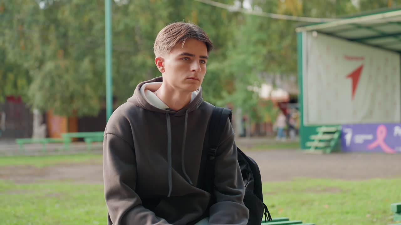 adolescente branco sentado em um banco com uma mochila, luz solar suave filtrando pelas árvores, expressão calma e contemplativa, coreto do parque no quadro, gradação de cores cinematográfica, clima de planejamento tranquilo e sonhos futuros