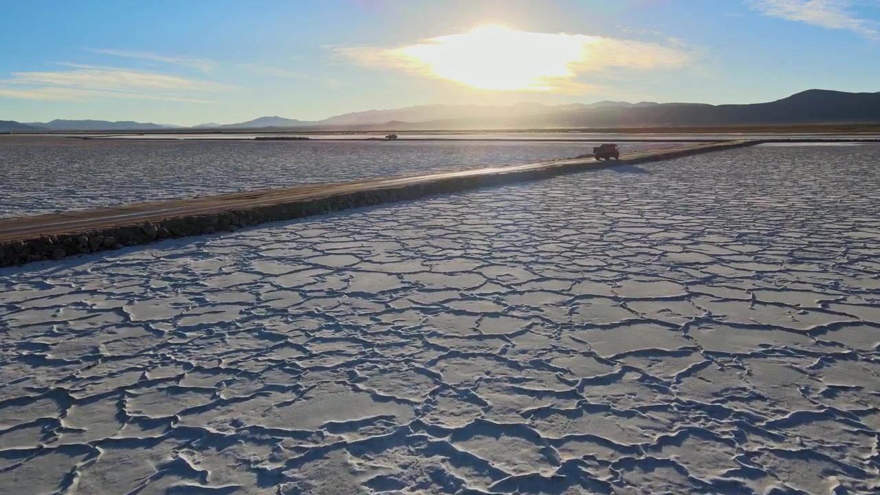 Vehicles drive along a narrow raised road cutting through patterned Salinas Grandes salt flats at sunset in northern Argentina.