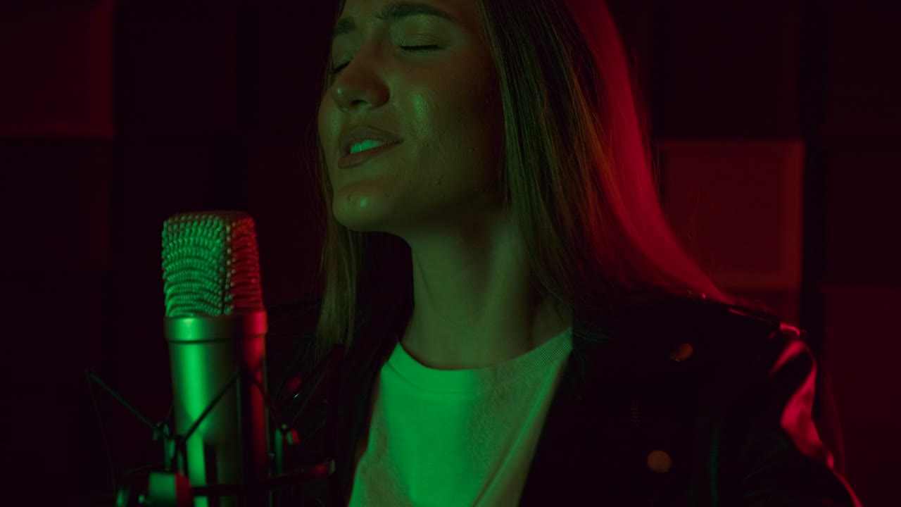 Attractive woman in glasses and headphones sings into a microphone in neon light in the dark studio. Emotional girl is recording a song on a black background