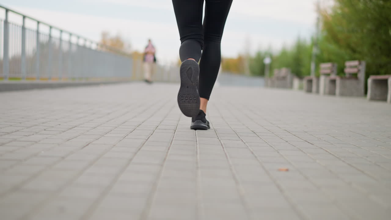 Close-up view of athlete woman running on pavement in black leggings and sneakers, with two women passing in blurred background, promoting active lifestyle and fitness
