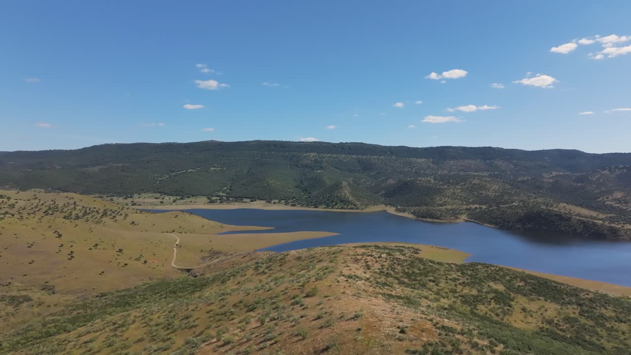Low forward flight over golden hills and green shrubs around the Cijara reservoir, full from spring rains. Blue-green water reflects sky and forests under a sunny May sky in Badajoz, Spain
