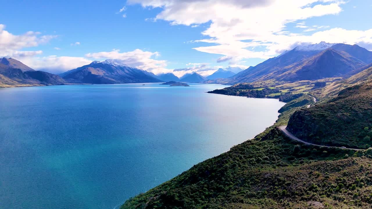 Drone glides above winding road, lush hills, and blue lake under bright daylight, wide perspective
