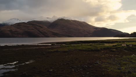 Aerial-drone-footage-reversing-over-the-shore-of-Loch-Etive-in-Glen-Etive-in-the-Highlands-of-Scotland-in-winter-to-reveal-a-snow-capped-mountain-at-sunset-with-golden-clouds