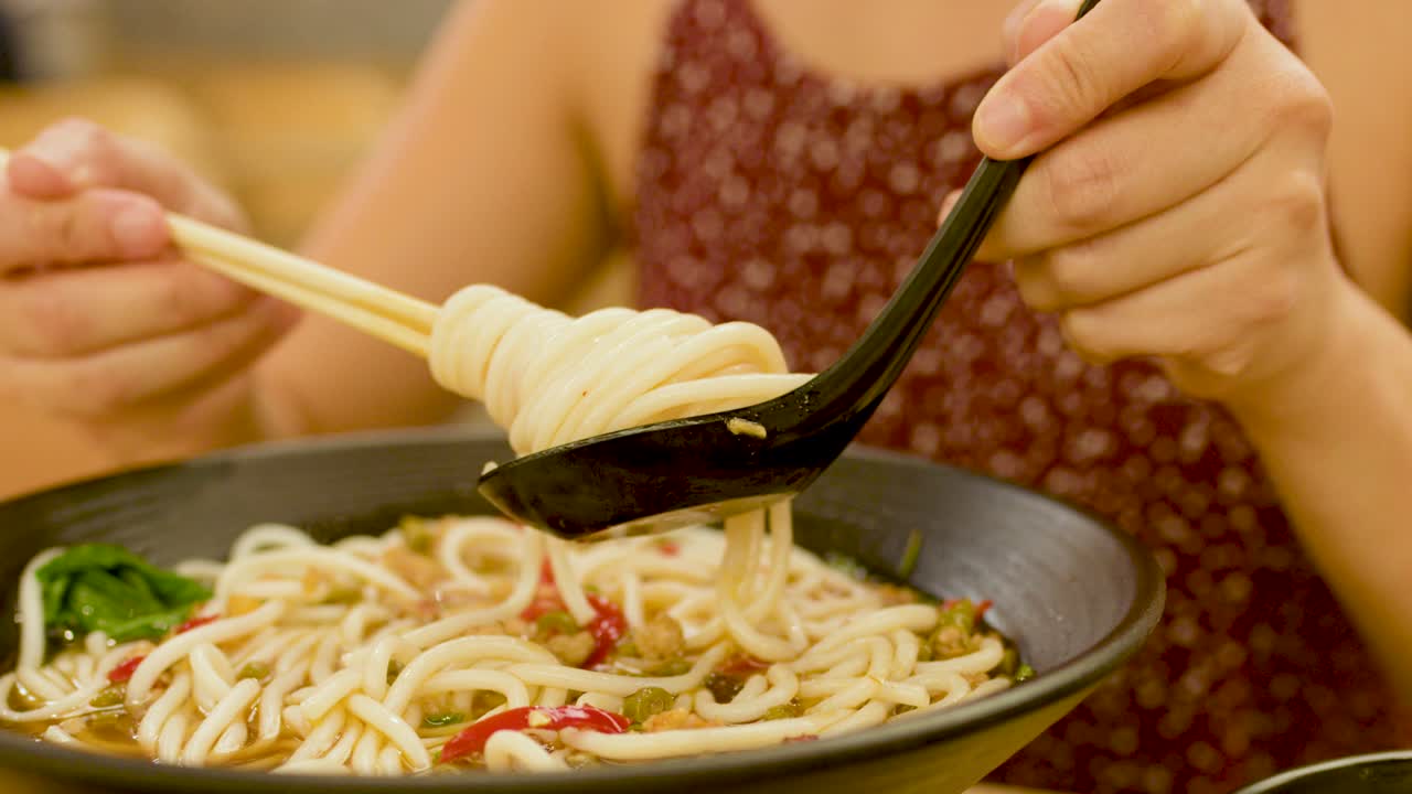 Close-up of woman using chopsticks and spoon to lift noodles from spicy Asian soup bowl