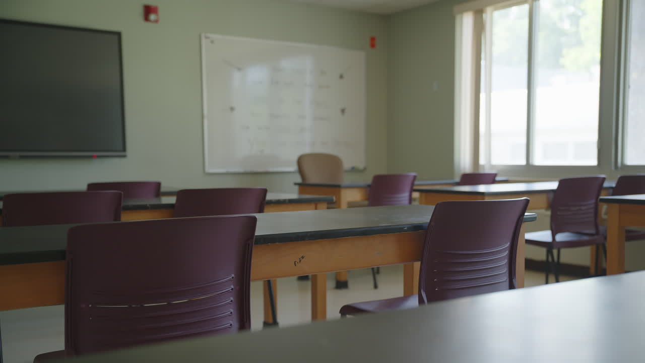 Daylit school classroom with maroon chairs, wood desks, whiteboard, and flat screen monitor