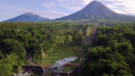 aerial backwards flight showing natural lake surrounded by wilderness of indonesia and giant merapi volcano in background during summer - bego pendem,indonesia