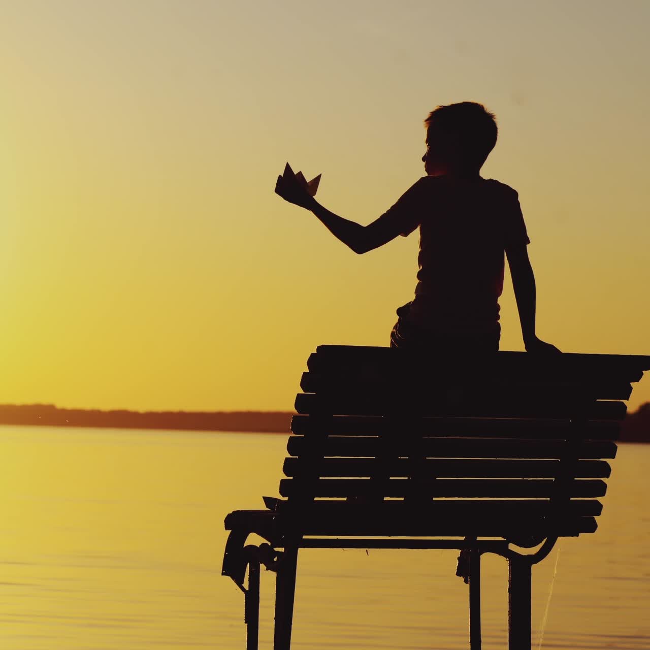 Little boy is playing with a paper boat on an old bridge by the river. Silhouette of little boy sitting alone at the river on sunset background