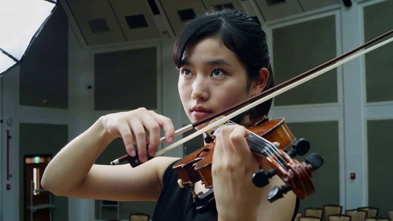 After conductor cue, violinist in black top raising violin, drawing bow across strings in rehearsal