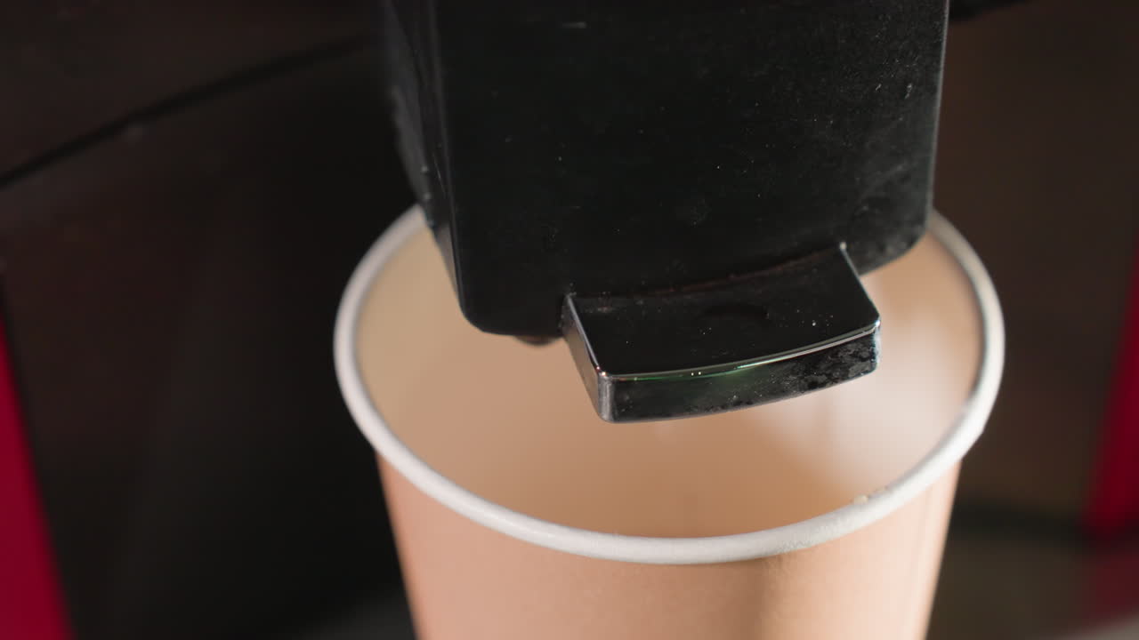 Close-up of espresso machine nozzle positioned just above a paper cup, with coffee being dispensed. Focus is on the machine's action as it fills the cup, demonstrating a caf -style coffee-making process