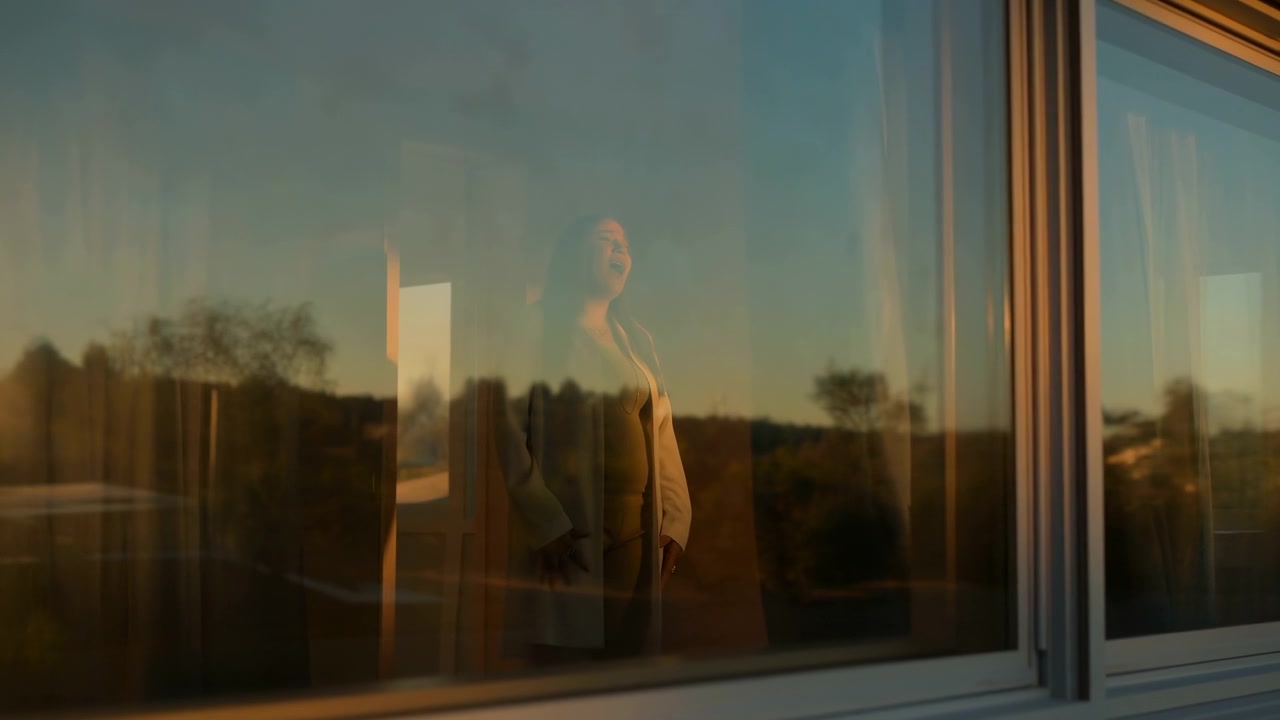暖かい夕焼けの中、現代的な家の中で幅広い窓のそばに立つ女性の姿。反射するガラスが室内のシルエットと融合し、柔らかな室内の色調と遠くの木立がその向こうに見える。
