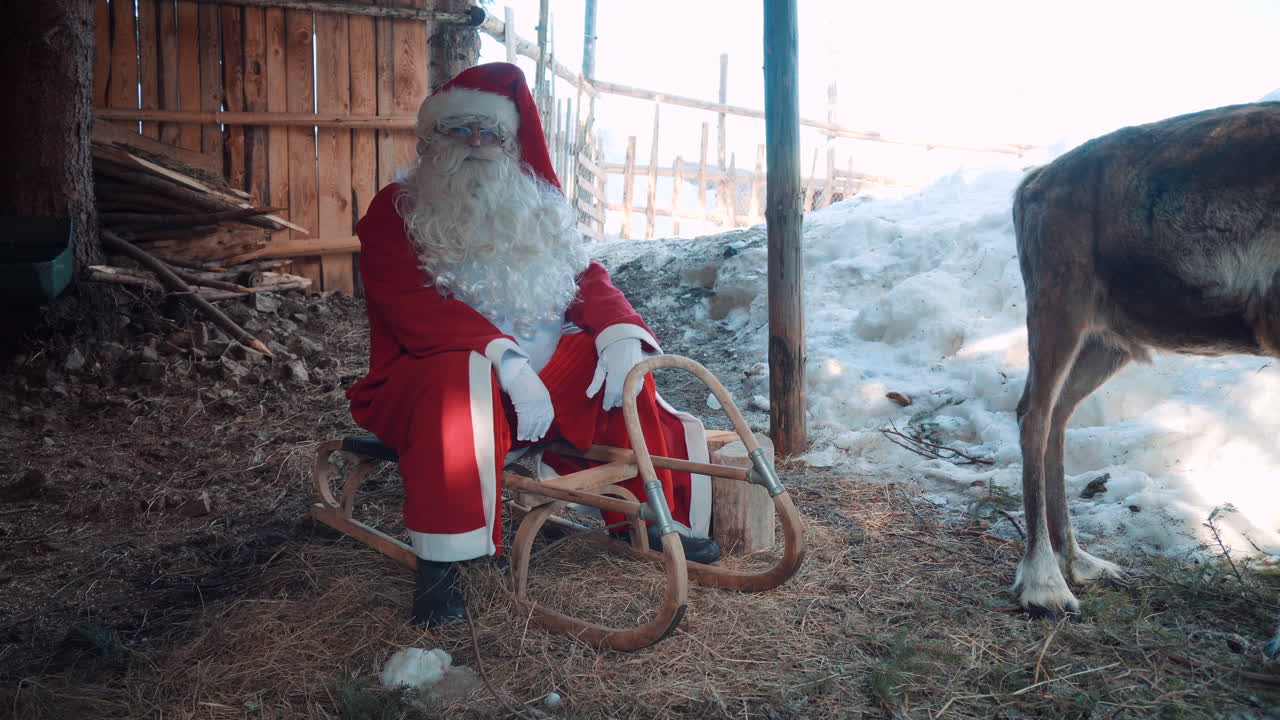 Babbo Natale all'aperto vicino a una casa di legno, seduto sulla sua slitta con una delle sue renne e che parla.