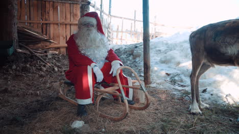 santa claus outside by a wooden house, sitting on his sleigh with one of his deer and talking