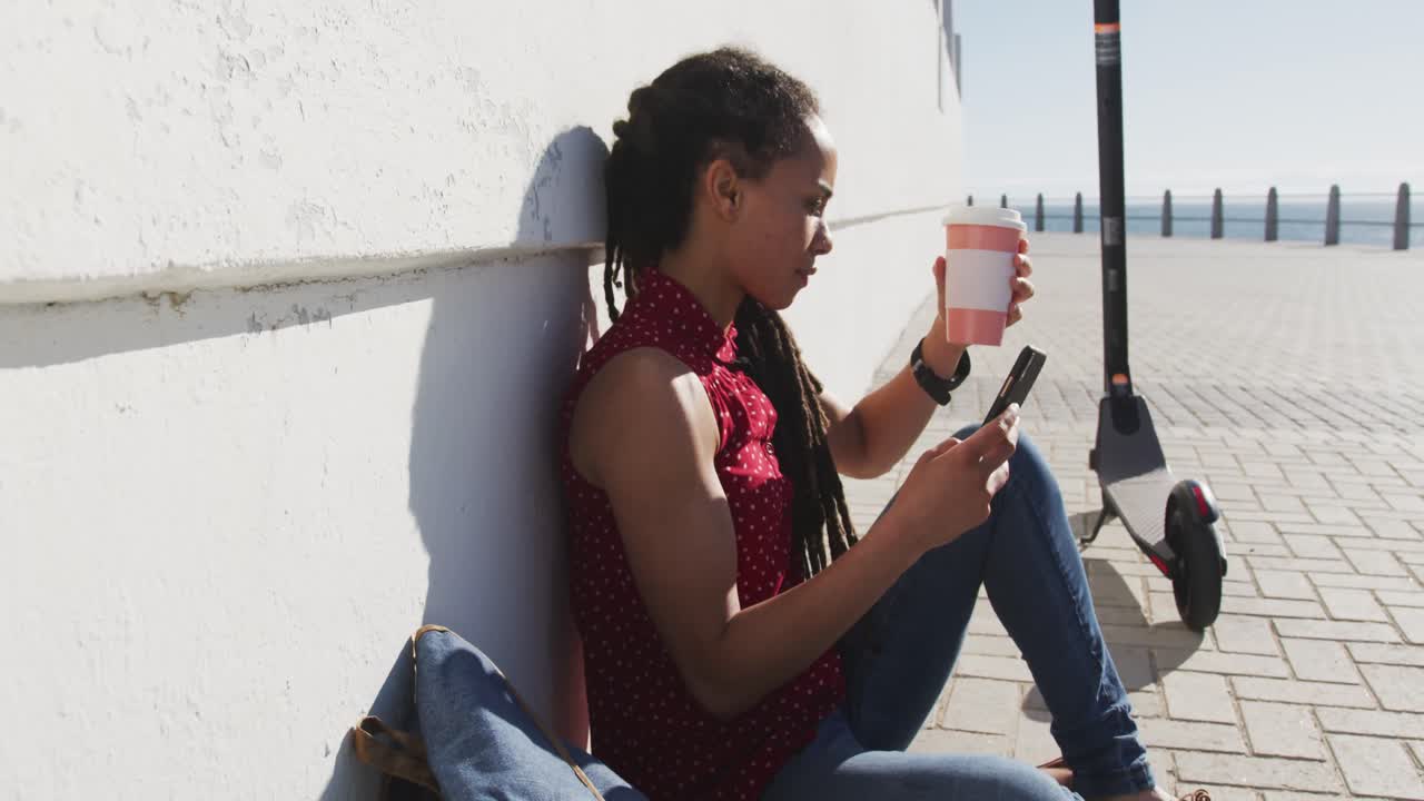 Donna afroamericana seduta che beve caffè e usa lo smartphone sul lungomare.