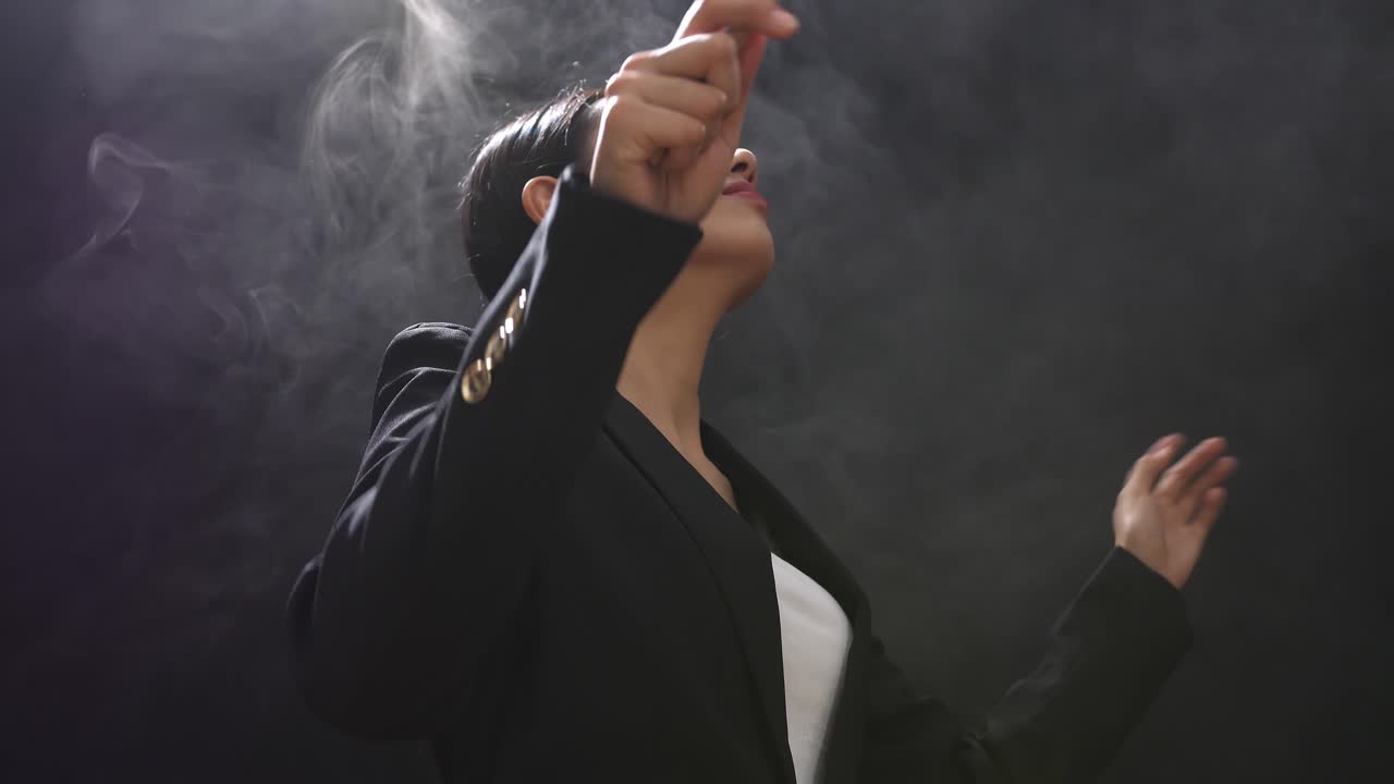 안개가 가득한 검은 스튜디오에서 지이를 들고 제스처를 빠르게 보여주는 아시아 지휘자 여성의 측면 시각