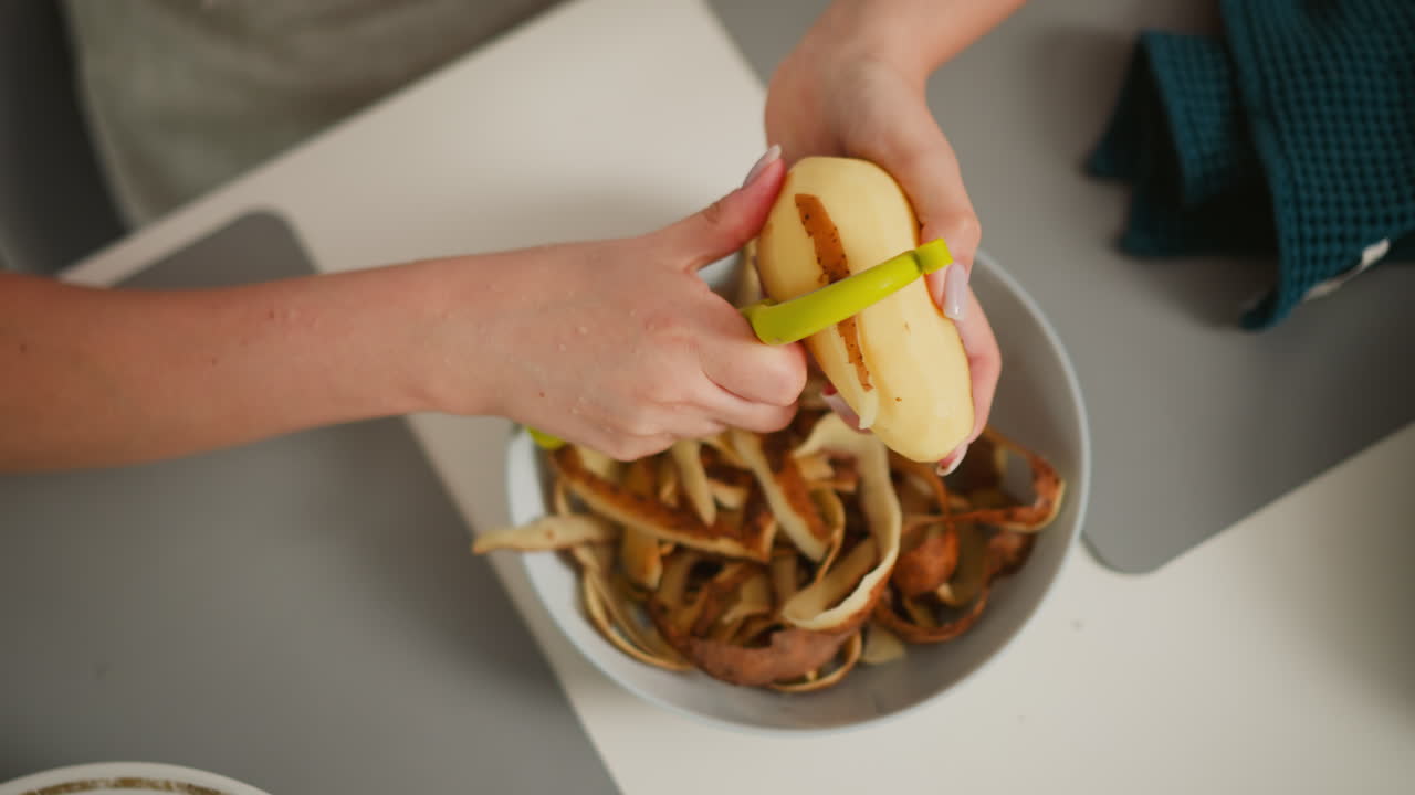 Overhead view of fair skinned woman peeling potato with green peeler over bowl filled with brown skins, kitchen cloth beside plate, focused hands removing final peel in clean cooking environment