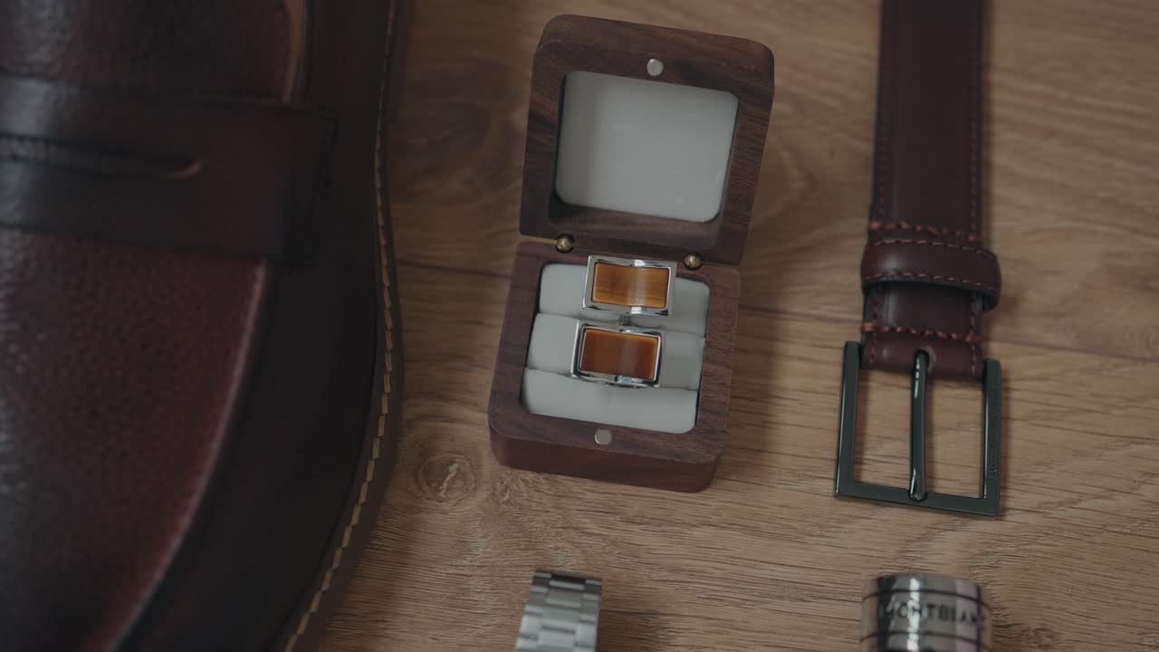 Flat lay of men’s wedding accessories on wooden surface including brown shoes, cufflinks, watch, and leather belt
