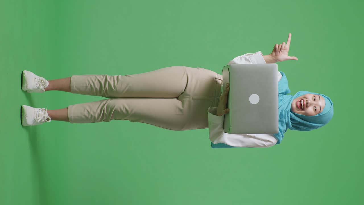 cuerpo lleno de mujer musulmana asiática sonriendo y escribiendo en una computadora portátil luego apuntando a un lado mientras está de pie en el estudio de fondo de la pantalla verde