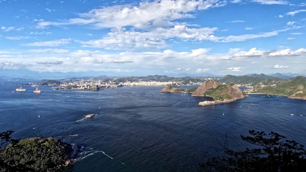 Time-lapse aéreo de Niterói em um dia ensolarado. Captura a vibrante Baía de Guanabara azul, montanhas tropicais exuberantes e o horizonte da cidade sob um céu claro com nuvens brancas em movimento