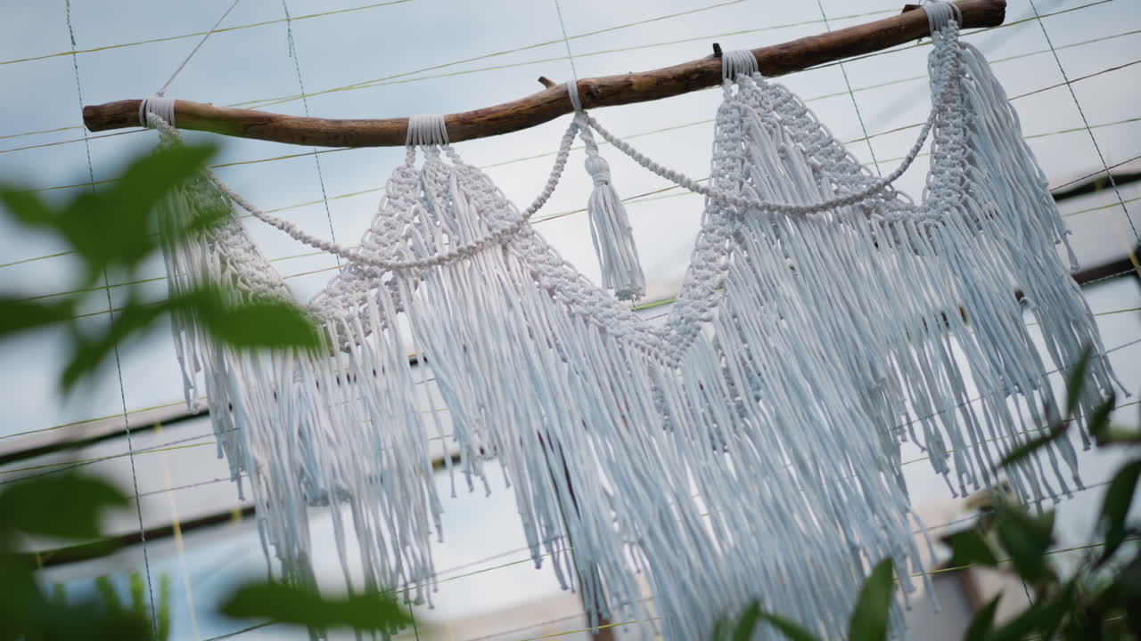 Móvil de macramé etéreo balanceándose sobre plantas, una barra de madera flotante que suspende largos flecos sobre una rejilla de invernadero, hojas flotando en primer plano, una instalación estilizada que captura un ambiente botánico tranquilo con luz natural suave.