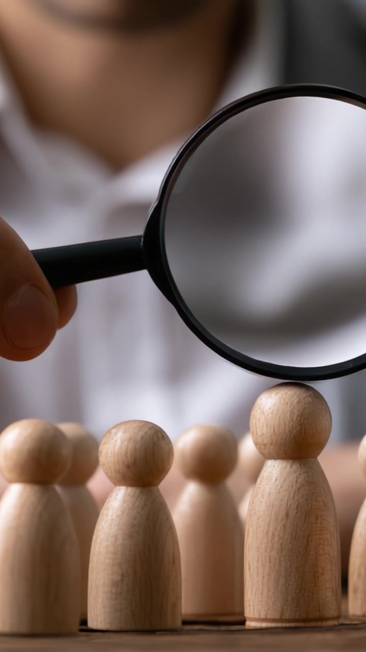 Focus op sleutelfiguren: een hand die een vergrootglas boven houten beeldjes houdt, wat analyse, observatie en selectie in een groepscontext symboliseert.
