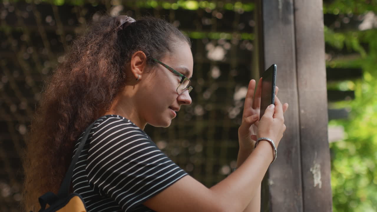 Young woman in striped shirt focused on smartphone near wooden post, capturing plant texture and light quiet contemplative mood, student researcher and amateur photographer roles, gentle bokeh