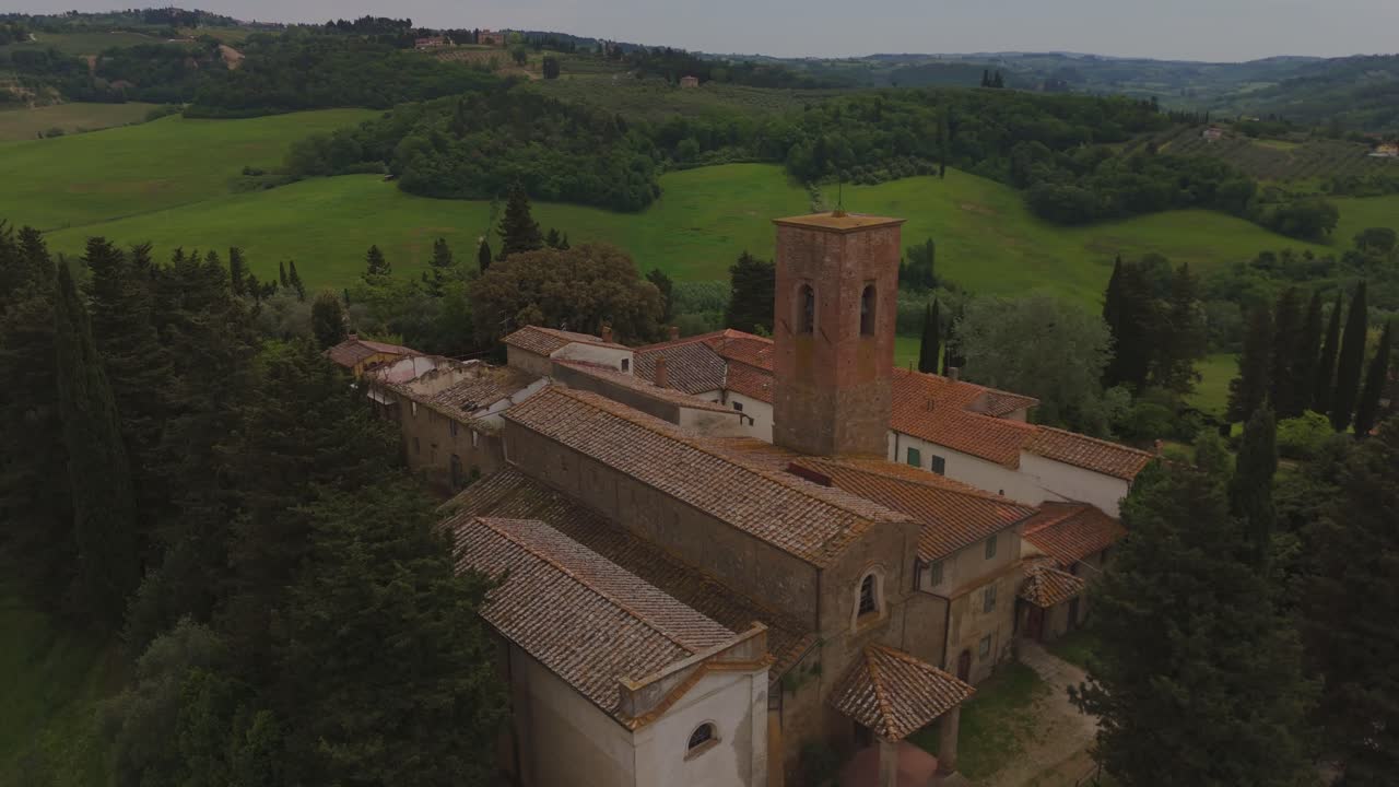Smooth orbit around a historic church in Tuscany, Italy. Surrounded by green hills, trees, and olive groves, the red-roofed structure with its medieval bell tower stands out in the rural landscape.
