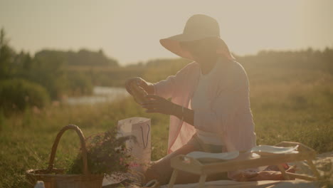 lady wearing sun hat sitting on picnic mat outdoors, retrieving snacks from paper bag to place on plates on picnic table, flowers in woven basket nearby, sunlit background with warm natural light