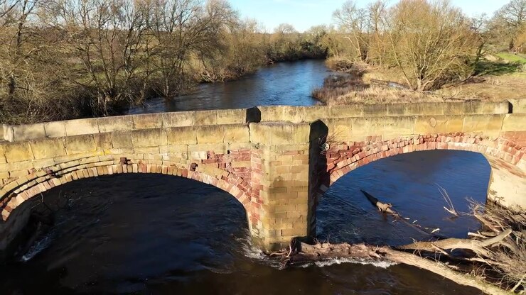 Ripresa aerea fluida con drone di un antico ponte ad arco in pietra su un fiume che scorre, circondato da alberi e dalla tranquilla campagna inglese nel Regno Unito