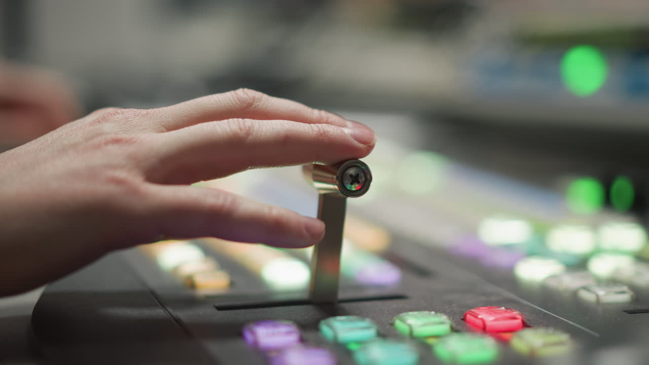 Close-up of hand adjusting broadcast control panel lever with colorful buttons, focusing on broadcasting and media production equipment in a television studio environment