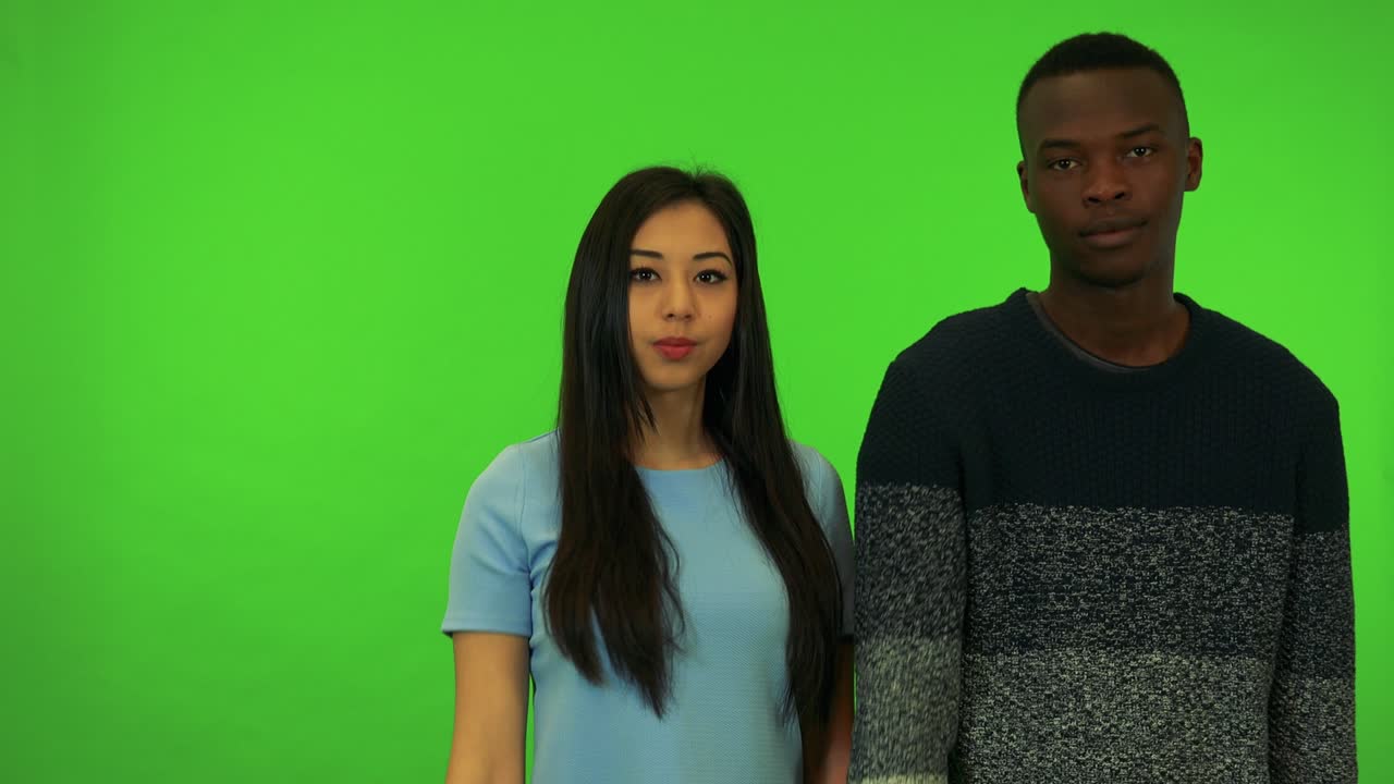 Um jovem homem negro e uma jovem mulher asiática mostram o polegar para baixo para a câmera - estúdio de tela verde