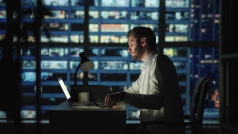 tired young man working on a laptop late night in the office. sleepy businessman sitting at desk in dark office. tired and stressed businessman in glasses works on a laptop of the night city office