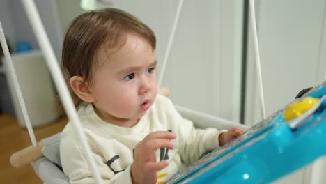 Cute-little-baby-sitting-in-an-indoor-doorway-swing-with-an-interactive-toy-board-surprised-to-glowing-button