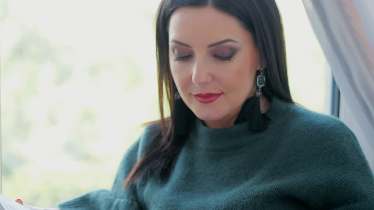 hermosa mujer encantadora con ropa verde relajándose en casa en el acogedor alféizar de la ventana y leyendo un libro. dama sonriendo. cámara lenta