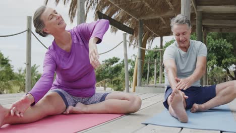 Happy-senior-caucasian-couple-practicing-yoga-sitting-on-beach-sun-deck,-in-slow-motion