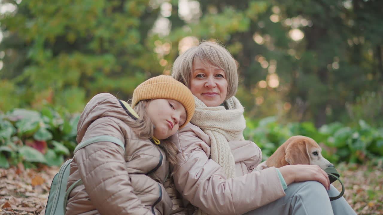 child leans sleepily on mom shoulder while mom smiles softly and holds dog close during peaceful moment in autumn forest with golden leaves scattered on ground and soft evening light