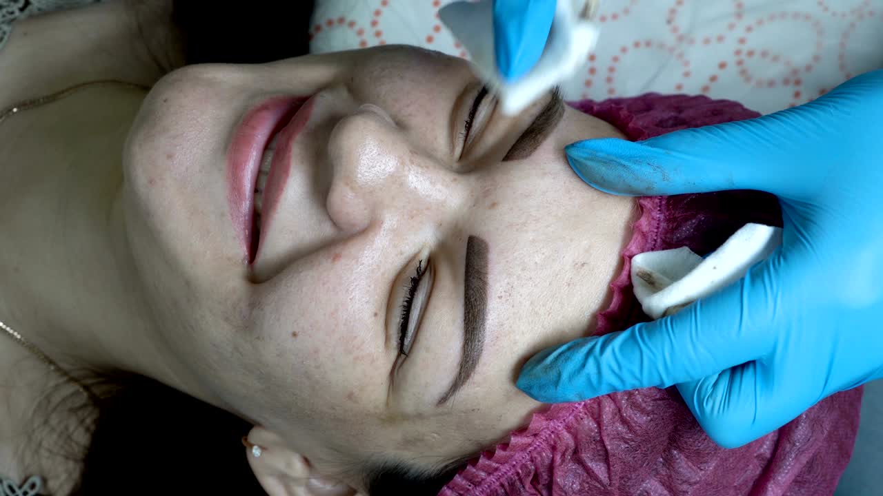 The beautician treats the skin around the eyebrows of the girl in the beauty salon after the procedure of permanent makeup eyebrows. Extremely close-up. The view from the top. 4K, 25 fps.