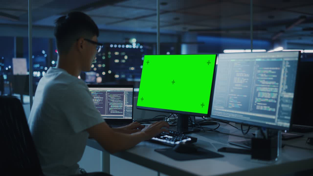 oficina nocturna: joven japonés trabajando en una pantalla verde chroma key computadora de escritorio. programador escribiendo código, creando software moderno, diseño de aplicaciones de comercio electrónico. por encima del hombro