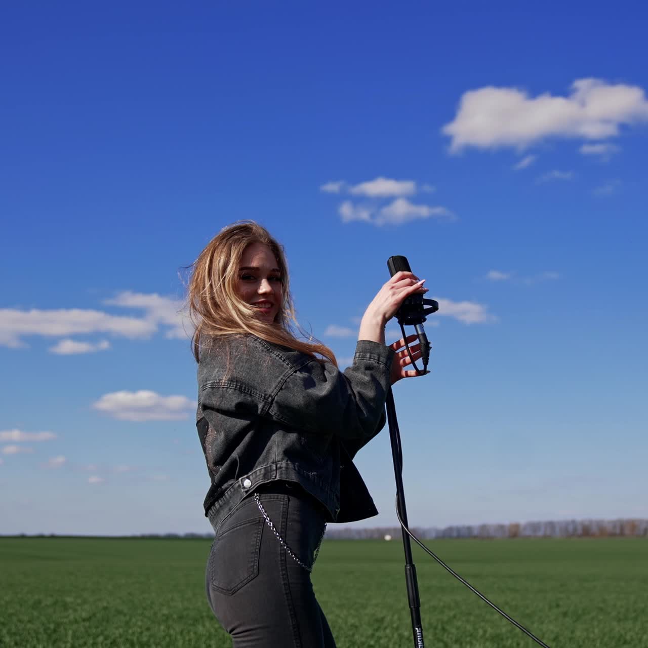Pop singer woman on sky background. Beautiful girl in jeans clothing dancing and singing into the microphone on field in sunny day