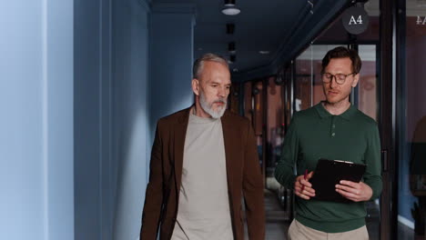 two businessmen walk and talk in an office hallway