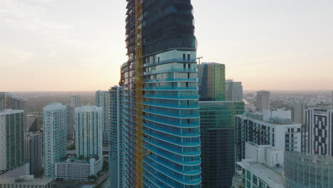 Fly-around-futuristic-tall-downtown-skyscraper-under-construction.-Revealing-modern-buildings-with-glass-facades.-View-against-sunset.-Miami,-USA