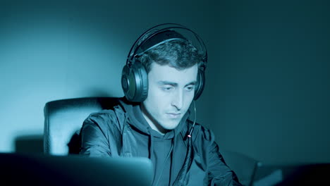 young caucasian hacker with headphones sitting at night in front of laptop screen, looking at camera and grinning