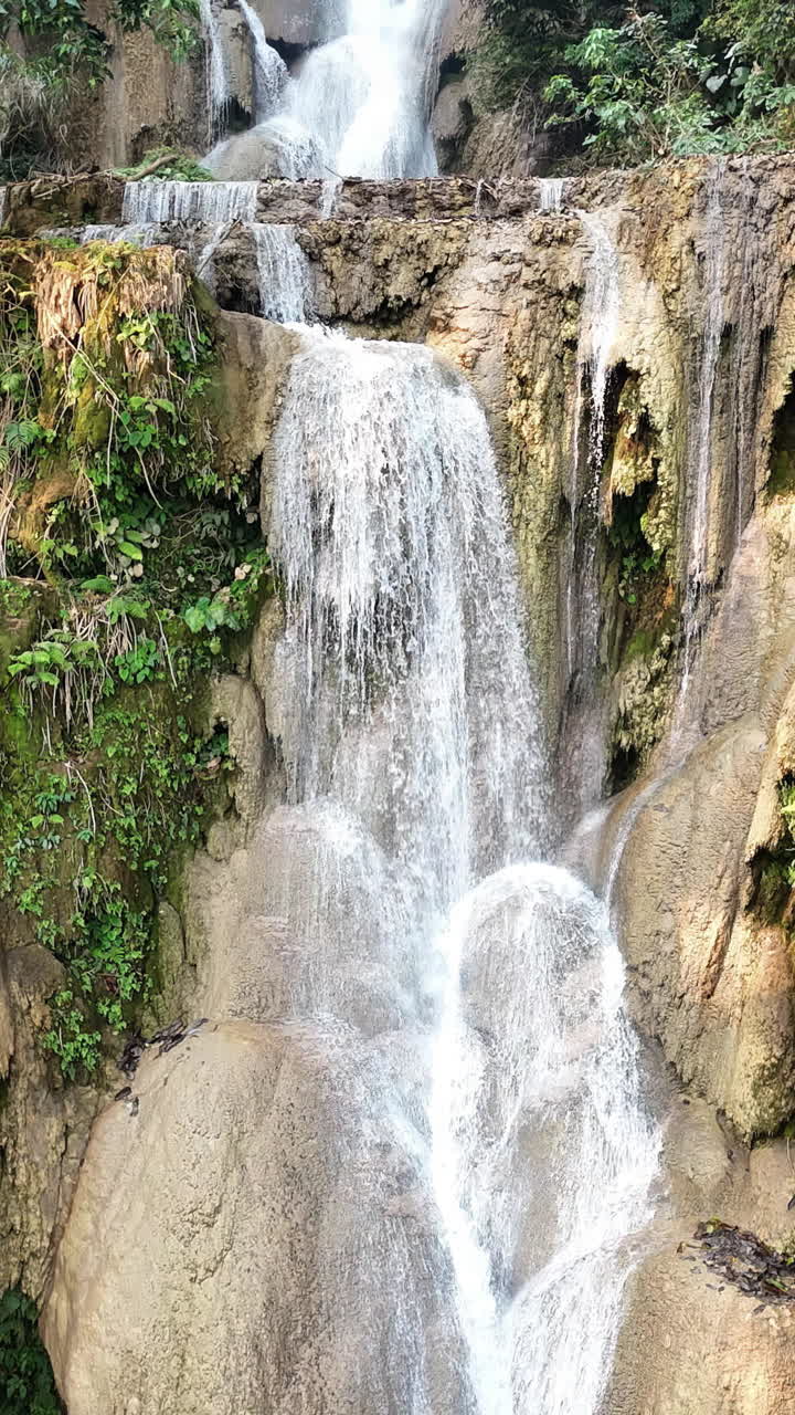 Vista aerea con drone della cascata principale di Kuang Si, vicino a Luang Prabang, dove numerosi corsi d'acqua scendono da formazioni rocciose calcaree in piscine turchesi circondate da una fitta giungla. Verticale