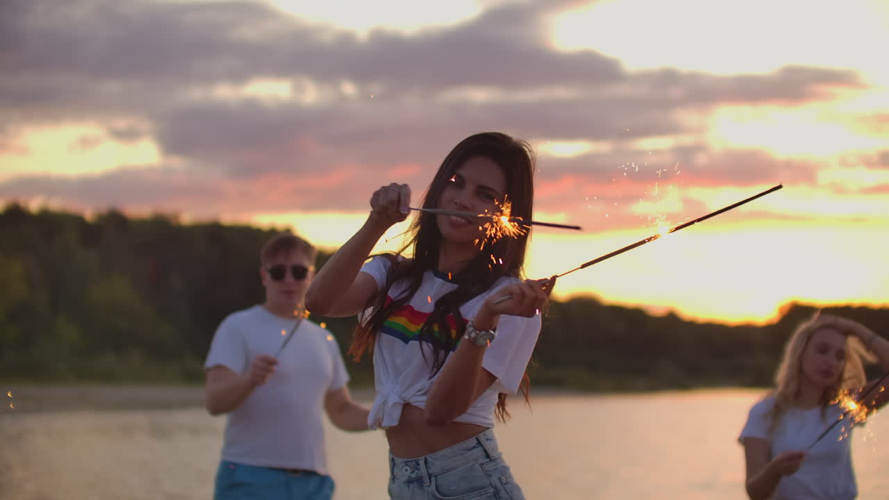 The female student with nude waist is moving her hands with big bengal lights on the sand beach with her friends. This is theamy summer evening on the open air party.