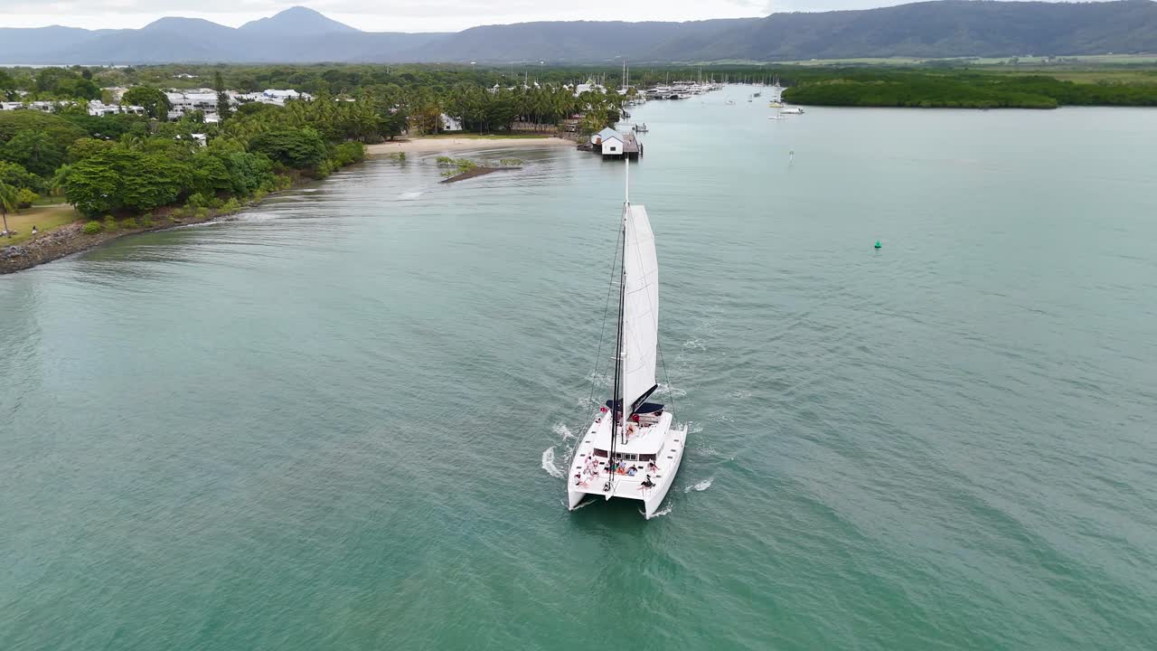 Drone captures a catamaran sailing through turquoise waters near lush greenery under overcast skies in Port Douglas, Australia