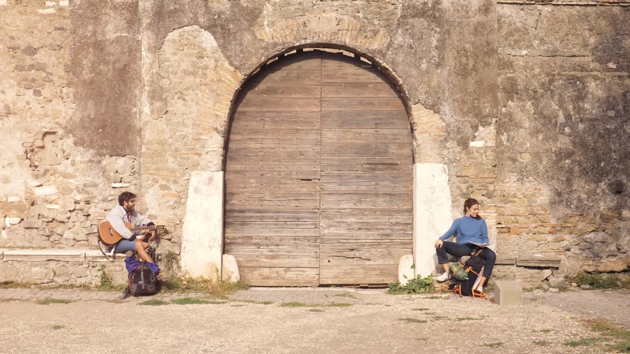 счастливая молодая пара туристов, сидящих возле деревянной двери средневекового здания в парке делли акедотти, руины парка в риме на рассвете с гитарой и картой.